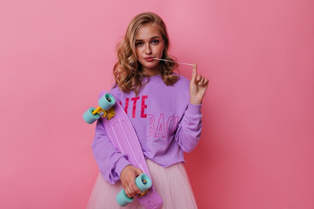 Spectacular white girl with curly hair posing on pastel. Glamorous female model with colorful skateboard.