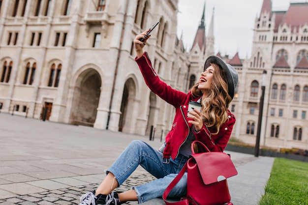 Free photo spectacular young woman sitting on the ground with backpack and taking picture of herself