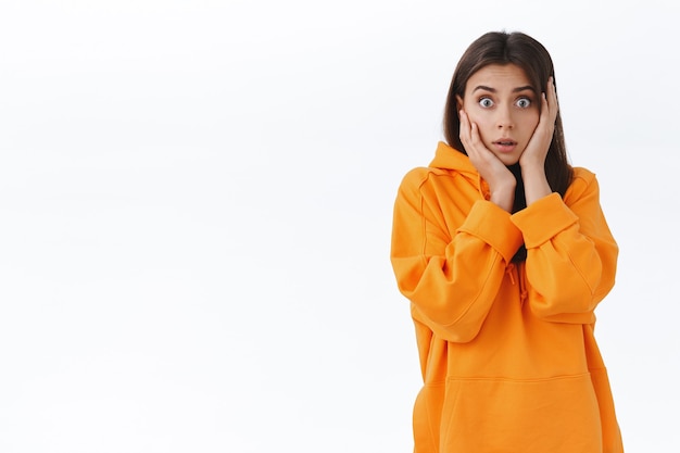 Speechless surprised and worried girl gasping stare camera startled, grab head open mouth, hear shocking unbelievable news, standing in orange hoodie concerned with rumor, white wall