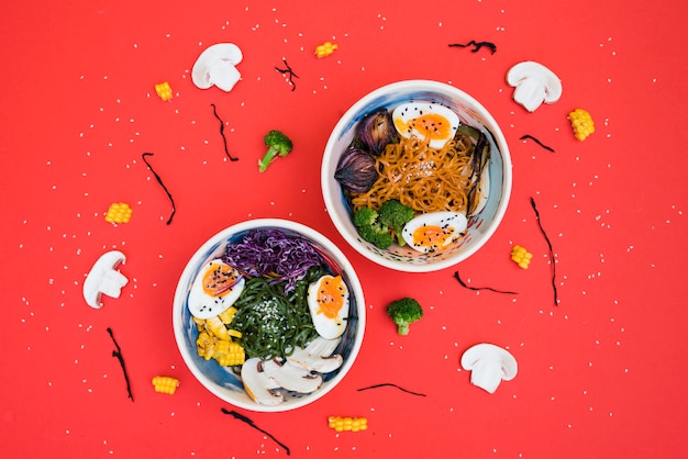 Free photo spicy ramen bowls with noodles; boiled egg and vegetables served with seaweed salad on red backdrop