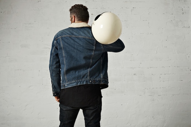 Spine view of biker wears shearling denim jacket and black blank henley shirt, holds vintage beige motorcycle helmet, isolated in center of white brick wall