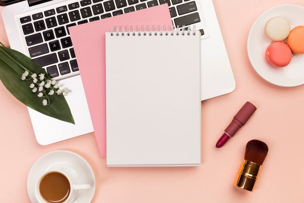 Spiral notepad on laptop with macaroons and coffee cup with makeup brushes on colored backdrop