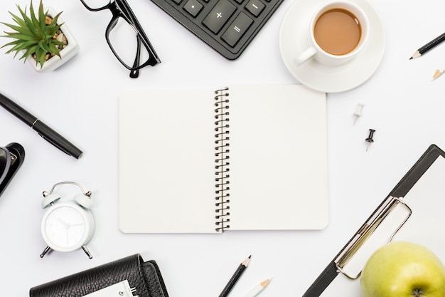 Spiral notepad surrounded with stationeries,apple and coffee on white office desk