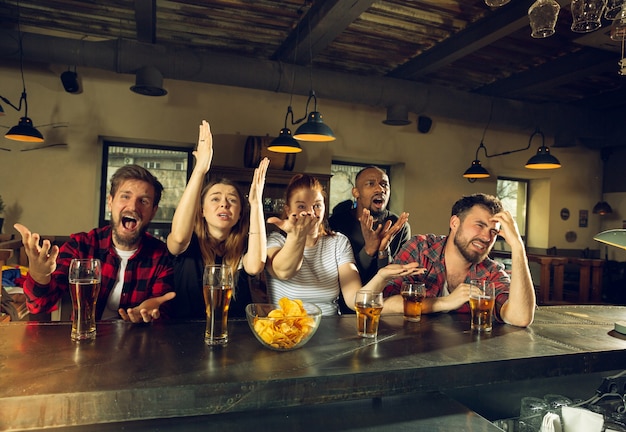 Sport fans cheering at bar, pub. Clinking beer glasses while watching championship, competition. Multiethnic group of friends excited in translation. Human emotions, expression, supporting concept.