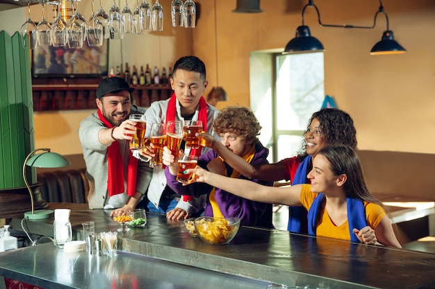 Sport fans cheering at bar, pub and drinking beer while championship, competition is going. Multiethnic group of friends excited watching translation. Human emotions, expression, supporting concept.