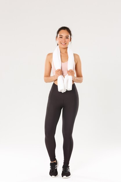 Sport, gym and healthy body concept. Full length of smiling cute slim girl, fitness trainer or sportswoman after exercises in gym, standing with towel wrapped around neck, white background.