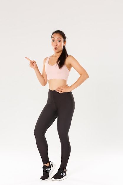 Sport, gym and healthy body concept. Full length of surprised cute brunette asian fitness girl in sportswear, looking amazed, pointing finger left, showing awesome prices for gym equipment.