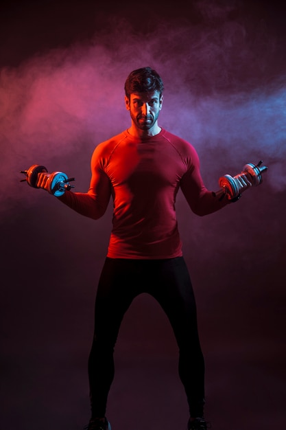 Free photo sportive man standing with dumbbells in dark