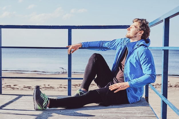 Free photo sportive young man is chilling near sea while sitting on balcony at bright sunny day.