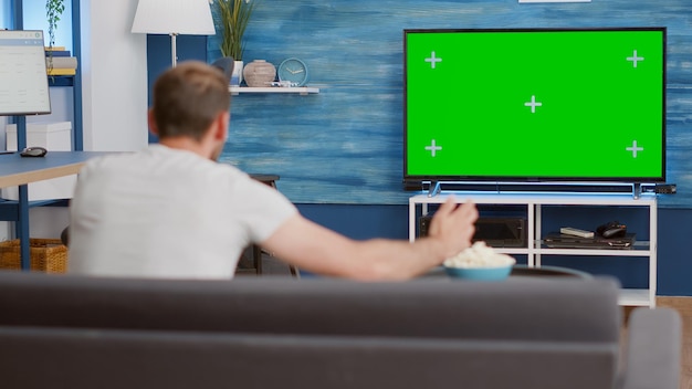 Free photo sports fan watching game on green screen tv mockup encouraging favourite team while relaxing at home sitting on couch. man sport supporter looking at television with chroma key display in living room.