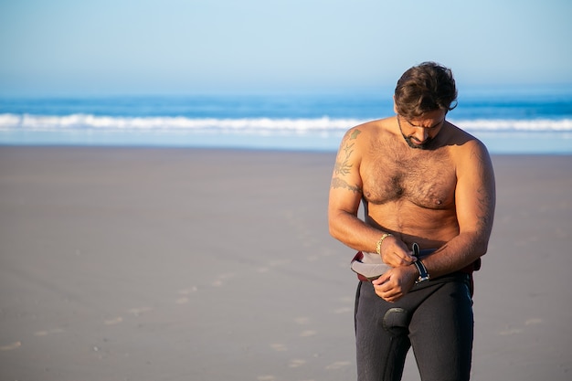 Sportsman putting on wetsuit for surfing on ocean beach and taking off watch