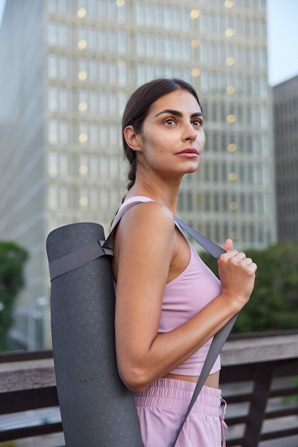 Free Photo  sportswoman in activewear carries rolled mat on shoulder going to have exercises in open air concentrated away poses on city scrapers