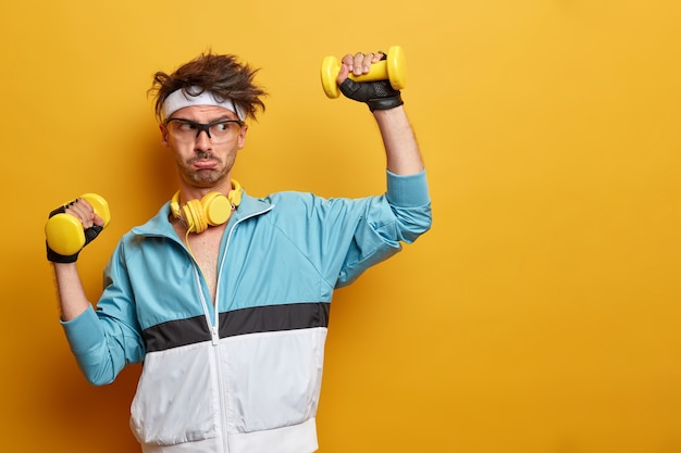 Free photo sporty athletic strong man lifts dumbbells and works hard on training biceps, leads active healthy lifestyle, has regular physical exercises, poses against yellow wall, empty space aside