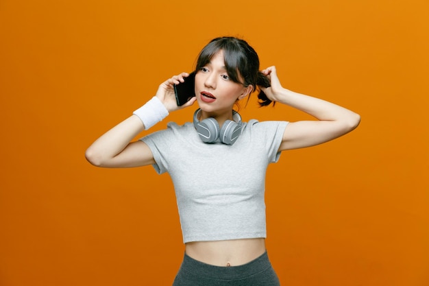 Sporty beautiful woman in sportswear with headphones talking on mobile phone looking confident and intrigued standing over orange background