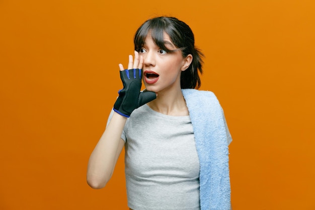 Sporty beautiful woman in sportswear with towel on her shoulder in gloves keeps hand near mouth like calling someone standing over orange background