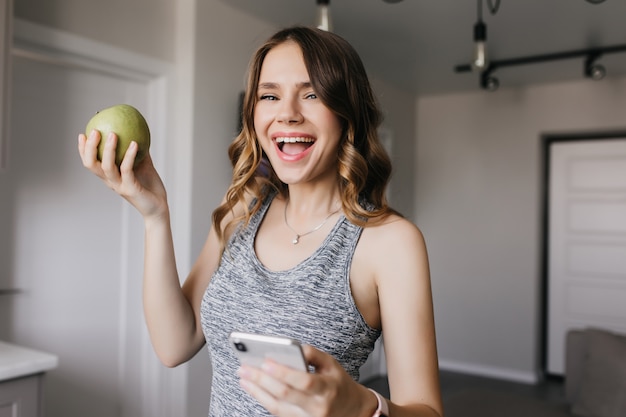 Sporty girl expressing happiness while posing with fruits. Indoor shot of relaxed curly woman with apple and smartphone.