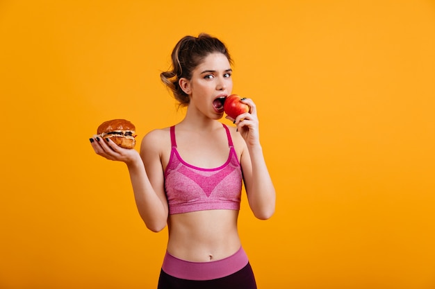 Free Photo sporty woman eats red apple on orange wall