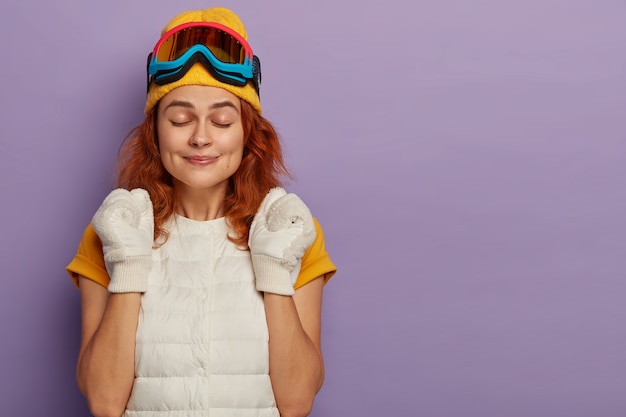 Sporty young woman enjoys ski resort, clenches fists with triumph, keeps eyes shut, wears protective snowboarding glasses, isolated over purple studio wall.