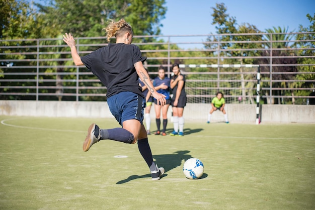 Sporty young woman kicking ball on summer day. Sportswoman in dark uniform kicking ball in direction of football goals, teammates in background. Sport, leisure, active lifestyle concept