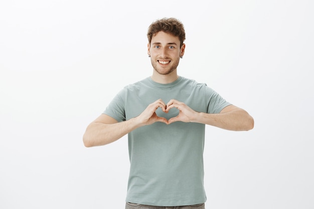 Spread love not war. Portrait of happy friendly-looking positive guy in earrings showing heart gesture over chest and smiling broadly while expressing affection or romantic emotions over gray wall