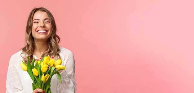 Spring happiness and celebration concept Closeup of happy and carefree blond european girl receive beautiful bouquet of flowers holding yellow tulips smiling and laughing pink background