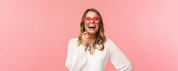 Free photo spring happiness and celebration concept closeup portrait of funny and carefree beautiful caucasian woman with blond hair white dress holding heartshaped glasses mask and laughing