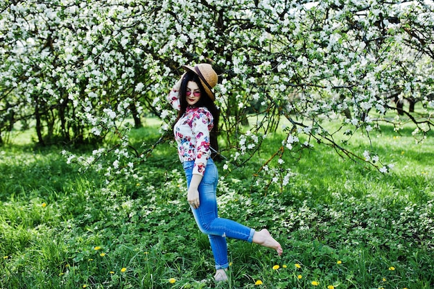 Spring portrait of brunette girl in pink glasses and hat at green blossom garden