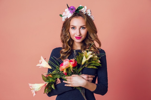 Free photo spring romantic  fashion portrait beautiful young female with long wavy blonde  hair in wreath of spring flowers  posing with flower bouquet over   pink  background.