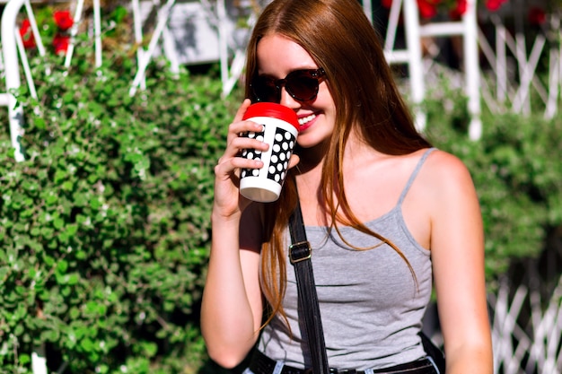 Spring summer positive portrait of young hipster girl, wearing street style casual outfit, walking at city center with cup of take away coffee, amazing natural ginger long hairs, tasty beverage.