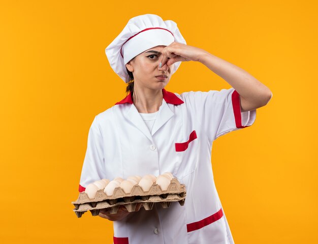 Squeamish young beautiful girl in chef uniform holding batch of eggs closed nose 