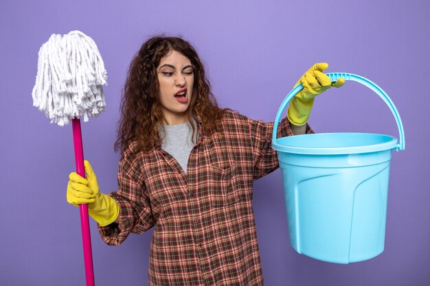 Free Photo squeamish young cleaning woman wearing gloves holding mop looking at bucket in her hand isolated on purple wall