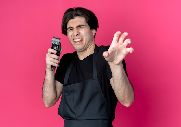 Free photo squeamish young handsome male barber in uniform holding hair clippers and showing tiger style gesture isolated on pink