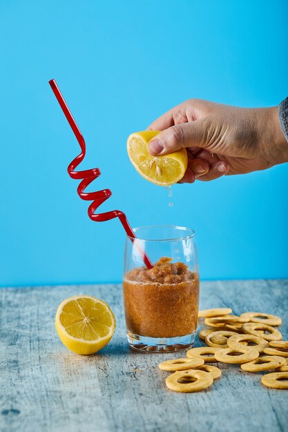 Squeezing fresh lemon into glass of juice with straw and crackers on gray table. 