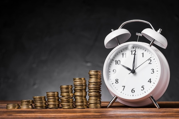 Free Photo stack of increasing coins with white alarm clock on wooden desk against black background