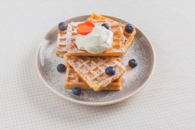 Free photo stack of waffles garnish with strawberry; blueberry and whipped cream on plate over the tablecloth