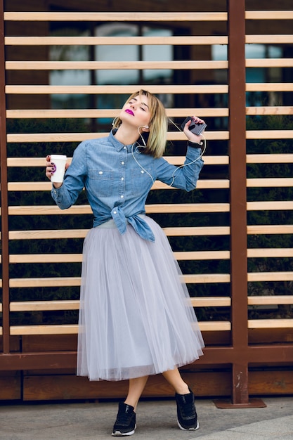 Free photo a standing blonde girl with pink lips holding a cup of coffee and listening to music on a smartphone with striped wooden balks behind