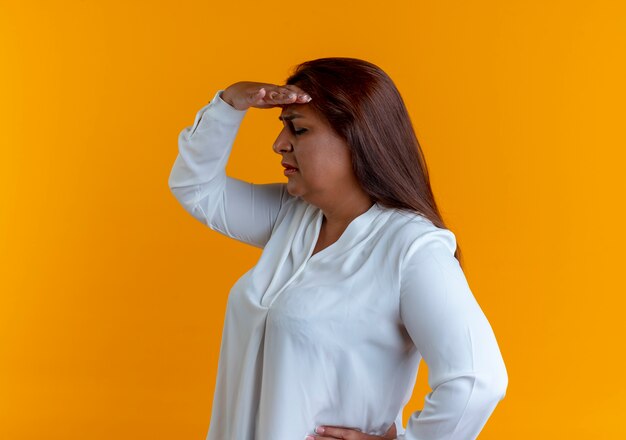 Standing in profile view casual caucasian middle-aged woman looking at distance with hand isolated on yellow wall