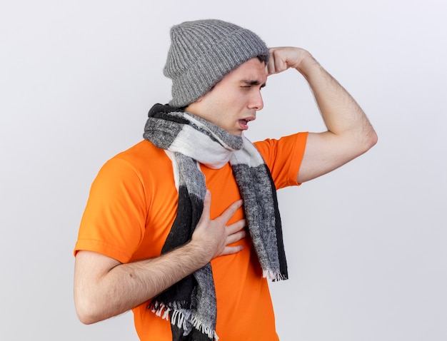 Free Photo standing in profile view young ill man wearing winter hat with scarf putting hands on forehead and aching chest isolated on white background