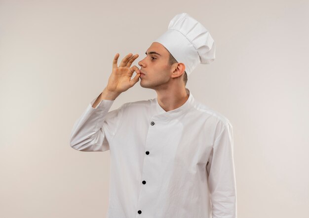 Standing in profile view young male cook wearing chef uniform showing delicious gesture with copy space