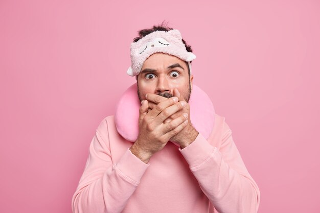 Startled amazed man covers mouth with frightened expression travels in transport for long distance wears sleepmask and travel pillow around neck 