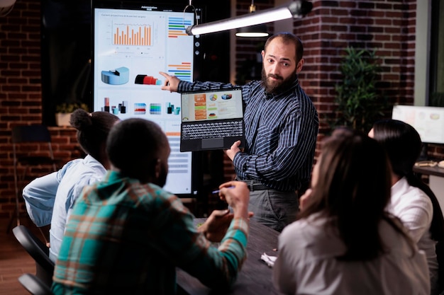 Free photo startup entrepreneur holding laptop with sales presentation charts pointing at wall screen tv in late night meeting. caucasian man presenting marketing strategy to coworkers working overtime.