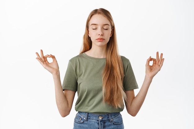 Free photo stay calm. relaxed young woman meditating, close eyes and hold hands sideways in zen mudra sign, practice yoga, breathing and resting her mind, standing over white background