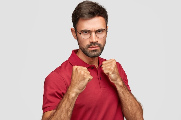 Free photo stern serious unshaven male keeps hands in fists, ready to fight with competitor, looks under eyebrows, has displeased expression, dressed in casual red t-shirt, stands against white wall indoor