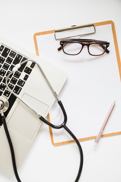 Free photo stethoscope over the laptop and clipboard with eyeglasses and pen on white background