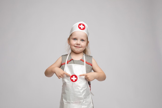 Free Photo stock photo of lovely little girl in white apron and a hat playing in a doctor she is pointing at her hat with white cross in red circle she is a doctor