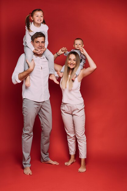 Stock photo of loving mother with baby son and dad with daughter sitting on shoulders and smiling happily on red background.