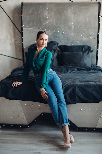 Free Photo stock photo portrait of attractive brunette woman in fashionable earrings, emerald blouse and jeans looking pensively away sitting on dark wooden floor against classic sofa.