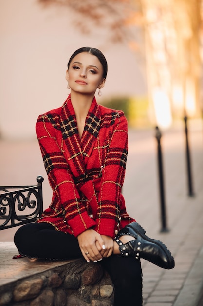 Free Photo stock photo portrait of attractive young girl in bright checked trench coat, black trousers and trendy boots sitting on bench in park. looking at camera dreamily.