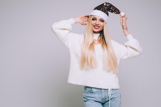 Free photo stock photo portrait of beautiful blonde woman in white sweater wearing bright sparkling santa hat posing on white background. copy space.