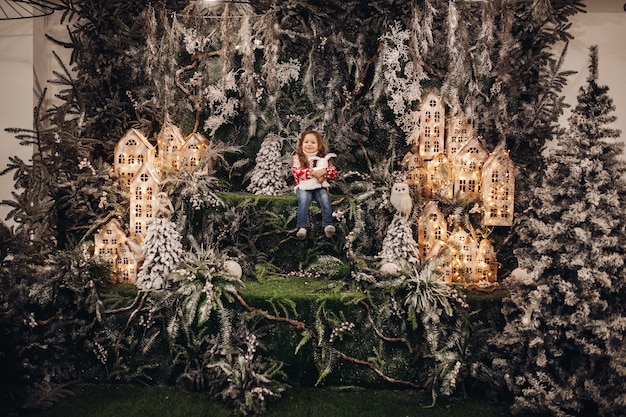 Stock photo of pretty brunette child in sweater and jeans embracing toy rabbit while sitting in beautiful Christmas decorations with illuminated houses and fir trees.